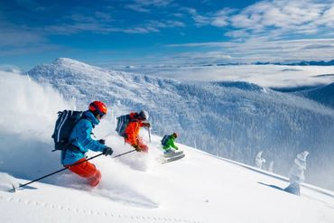 Three skiers descend a snowy mountain under a bright blue sky.