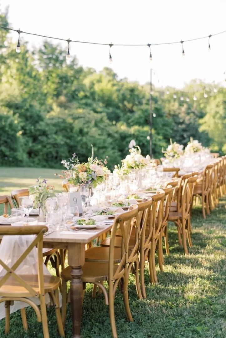 Elegant outdoor dining setup with wooden chairs and floral centerpieces on a long table.