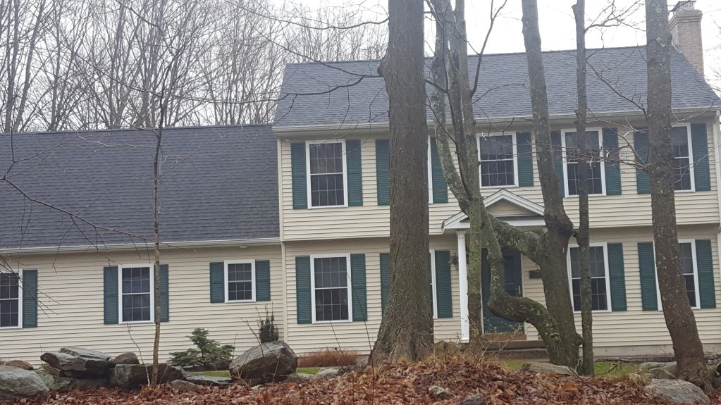 Large beige house with green shutters surrounded by leafless trees and rocks.