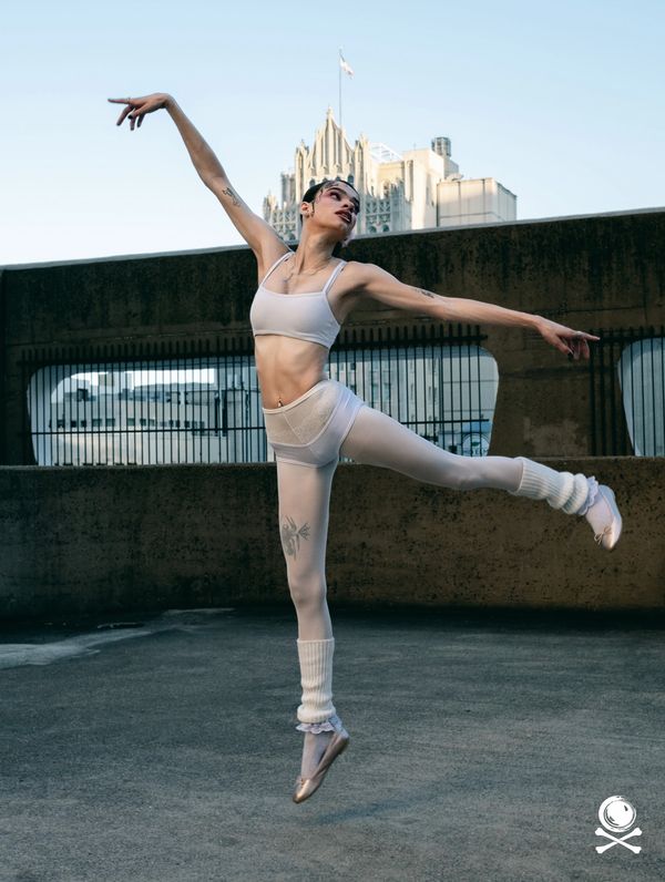 Ballet dancer in white attire gracefully leaps outdoors with cityscape backdrop.
