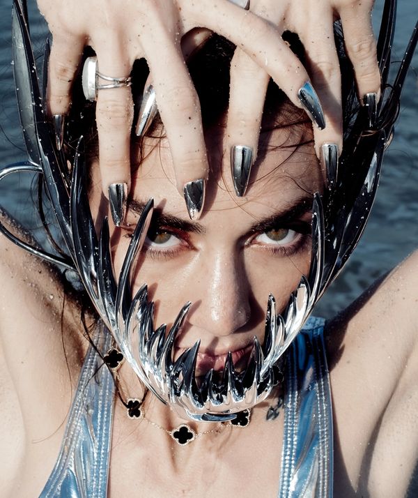 Close-up of a person with metallic claw-like nails and a shiny, spiked accessory framing their face.