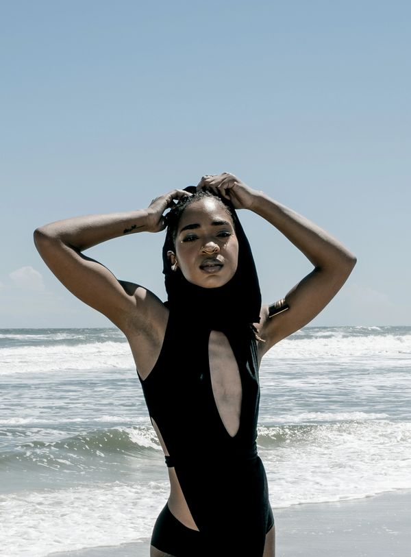Woman in a stylish black swimsuit posing at the beach with waves behind her.