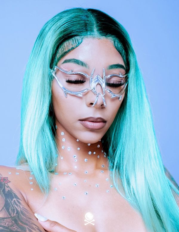 Woman with turquoise hair wearing unique silver glasses and rhinestones on neck and chest.