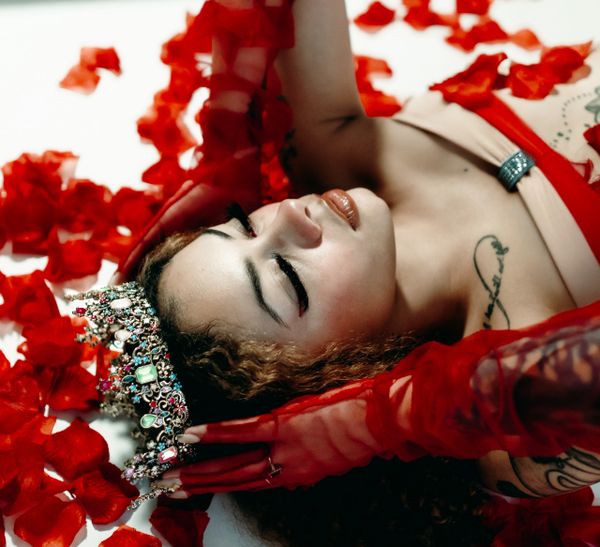 Woman wearing a jeweled crown and red gloves lies among red rose petals.