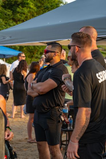 Security personnel in black shirts monitoring a crowded outdoor event on a sandy ground.