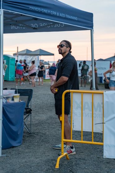 Man in sunglasses stands near a CPR training tent at an outdoor event.