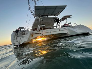 A catamaran named Edelweiss floats on calm water during sunset.