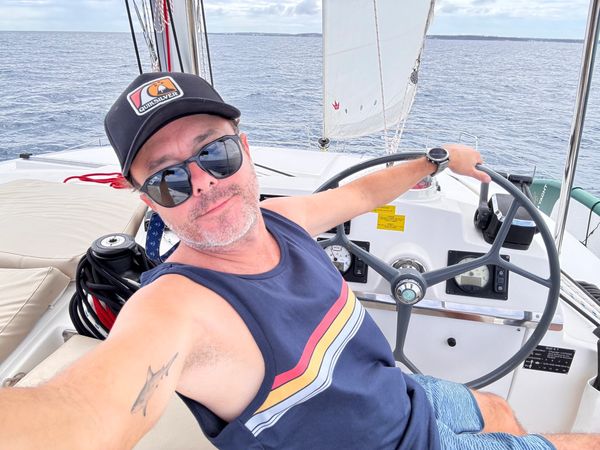 Man wearing sunglasses and a cap steering a sailboat on a calm sea.