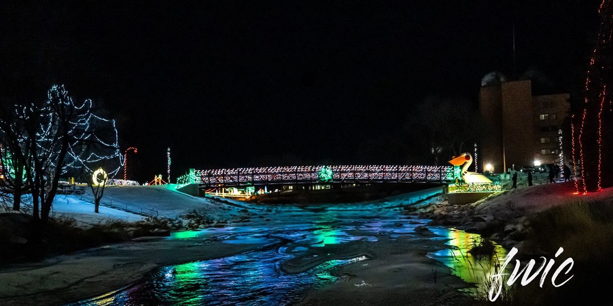 Bridge and trees decorated with colorful holiday lights reflecting on a frozen river at night.