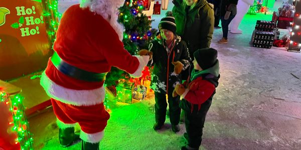 Santa Claus hands candy canes to children at a festive winter event with colorful lights.