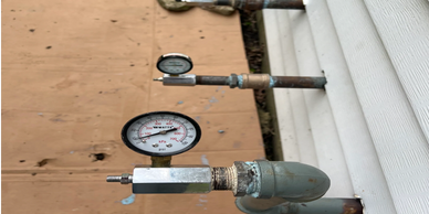 Outdoor pressure gauges attached to pipes by a white building.