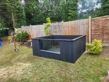 A black wooden raised garden bed with a glass window on the side in a backyard.