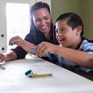 A woman happily helps a boy with Down syndrome draw at a table.