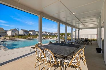 Covered patio with tables and chairs by a swimming pool in a residential area.