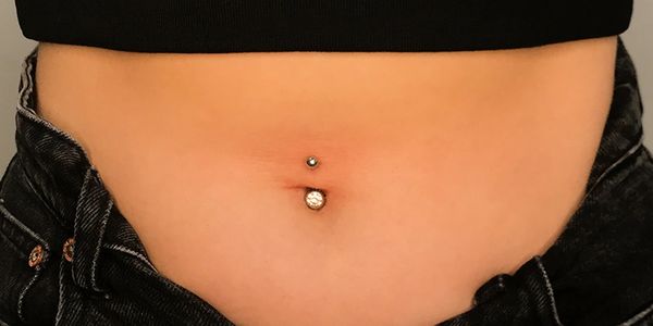 A photo of a person’s stomach with a silver barbell-style navel piercing featuring a small ball on t