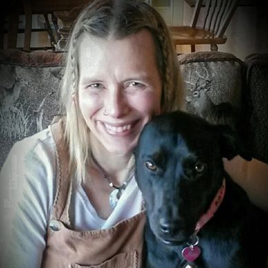 Smiling woman with braided hair posing with a black dog indoors.