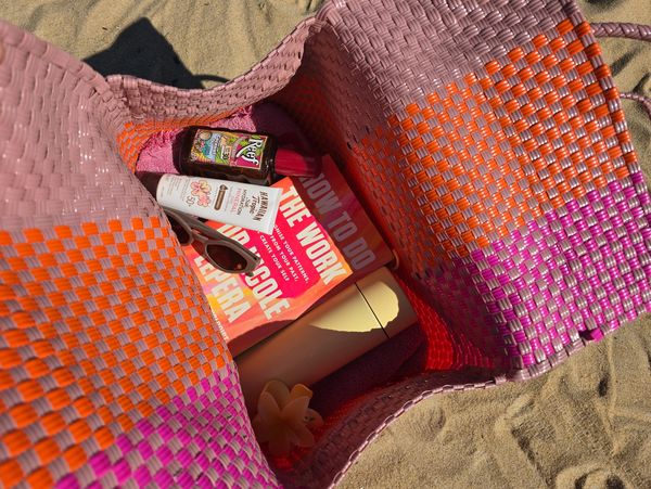 Open beach bag with sunscreen, sunglasses, a book, and a drink bottle on sand.