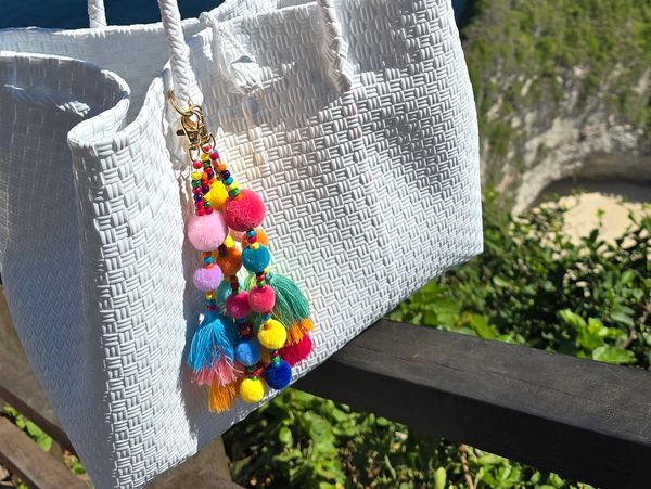 White woven handbag with colorful pom-pom keychains hanging outdoors.