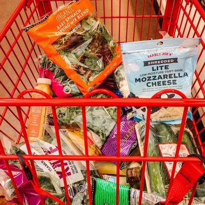 Grocery cart filled with food items including a bag of shredded cheese, bagged lettuce, dressing.