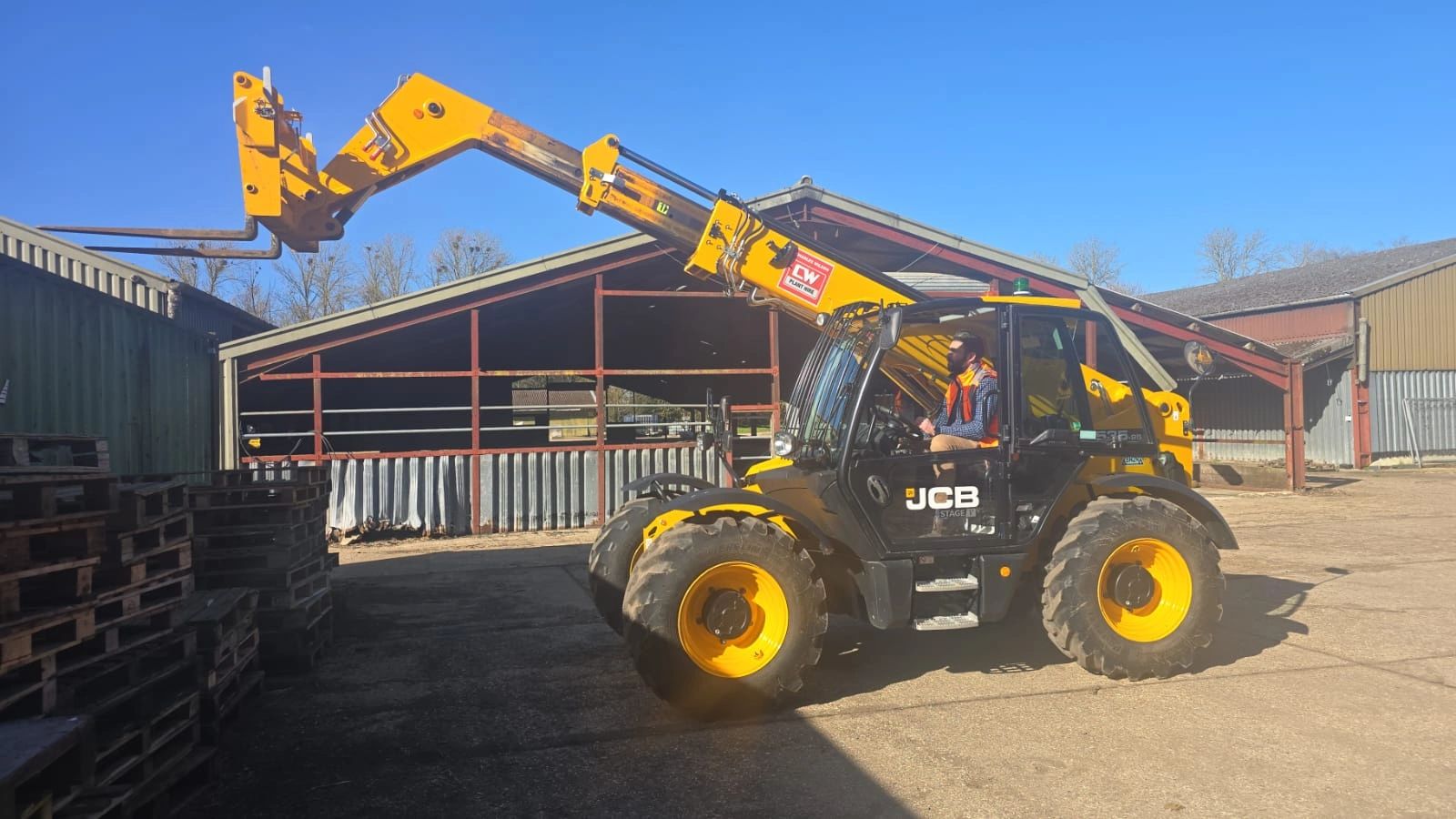 Yellow JCB telehandler lifting pallets near a warehouse on a sunny day.
