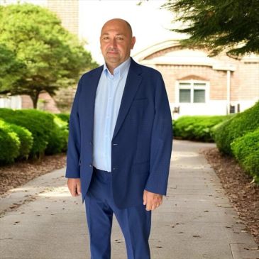 Man in blue suit standing on a sidewalk with greenery and buildings around.