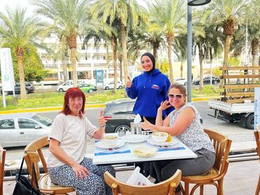 Three women smiling and giving thumbs up at an outdoor café with palm trees in the background.