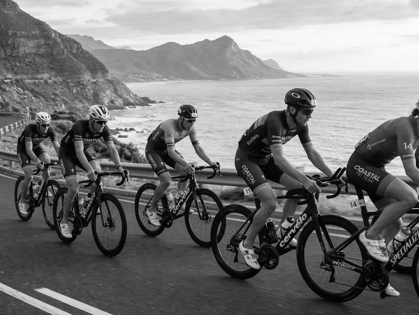 Cyclists riding along a coastal road with mountains and ocean in the background.