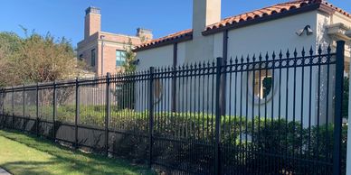 Black wrought iron fence surrounding a white building with bushes.