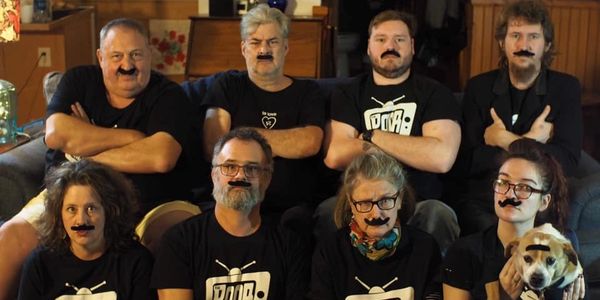 Group of eight people and a dog wearing fake mustaches indoors.