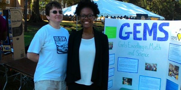 Two women stand in front of a GEMS science and math display poster outdoors.