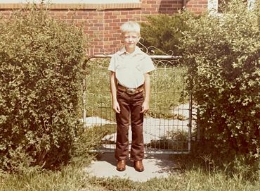 Young boy standing in front of a garden gate between bushes on a sunny day.