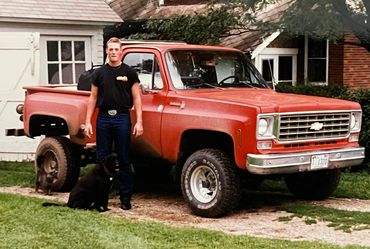 Man stands next to a red vintage Chevrolet truck and a black dog in front of a house.