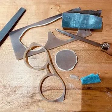 a buffalo horn frame cutout laying next to the raw material a scraper some sanding paper and a file