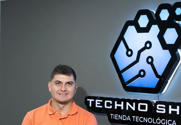 Man standing next to illuminated Techno Shop logo on wall.