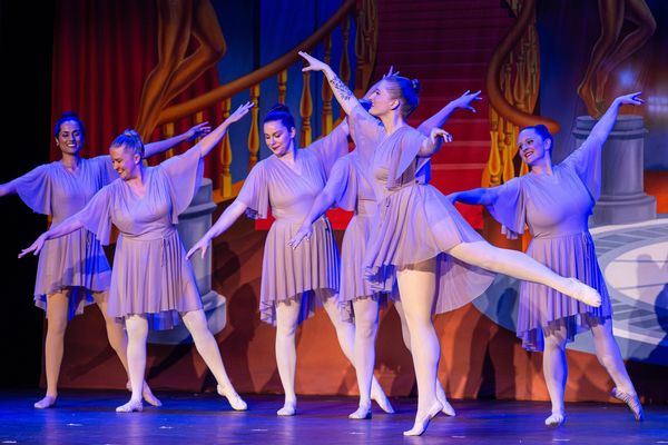 Six female ballet dancers perform gracefully on stage in matching lavender dresses.