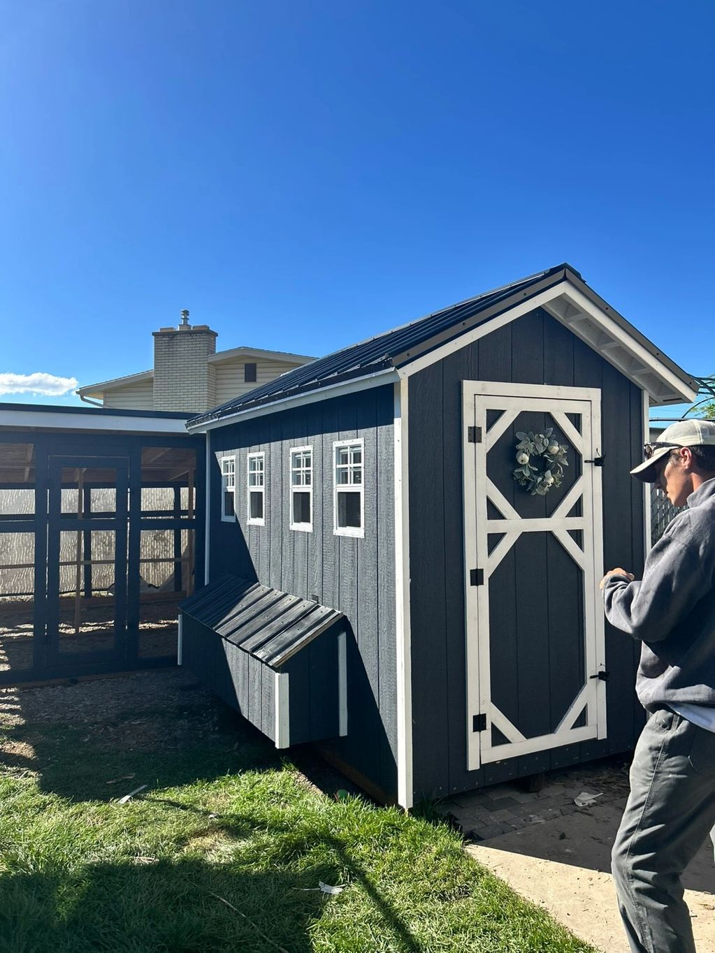 A man stands near a dark blue chicken coop with white trim under a clear blue sky.