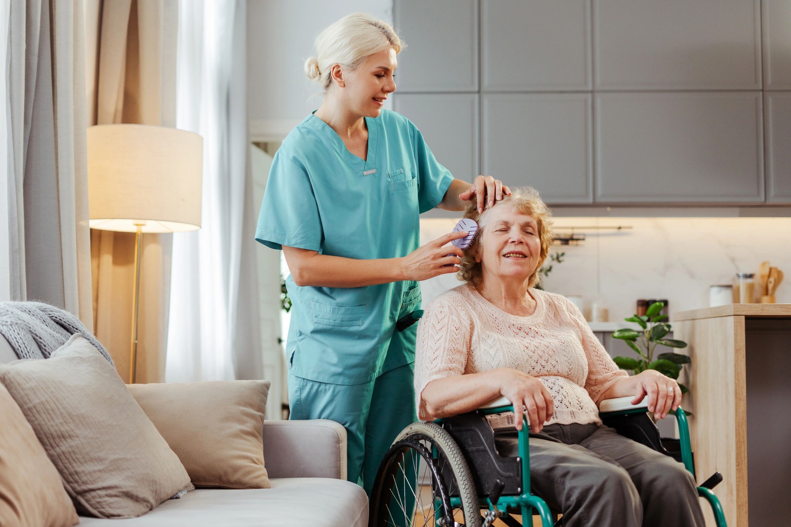 A smiling blonde caregiver brushes the hair of an elderly woman in a wheelchair.
