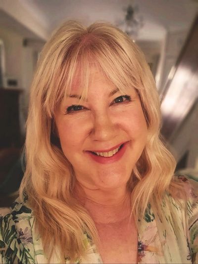 Smiling woman with blonde hair and floral top in a warmly lit room.