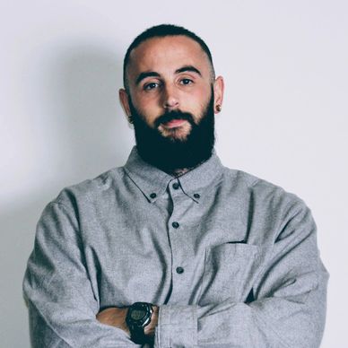 Man with a beard and earrings wearing a gray button-up shirt with arms crossed.