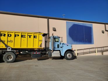 Yellow and blue waste disposal truck parked beside a beige building under clear blue sky.