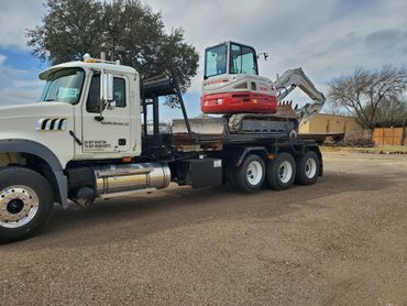A white truck carrying a red and white mini excavator on its flatbed.