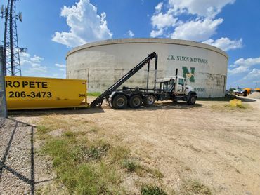 A truck with a dumpster in front of a large circular building under a blue sky.