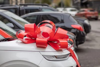 White car with a large red bow on the hood in a parking lot.