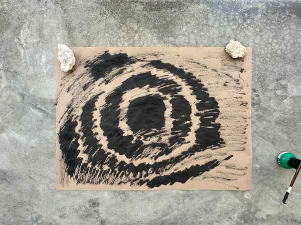 Abstract black ink spiral artwork on paper held by rocks on a concrete floor.