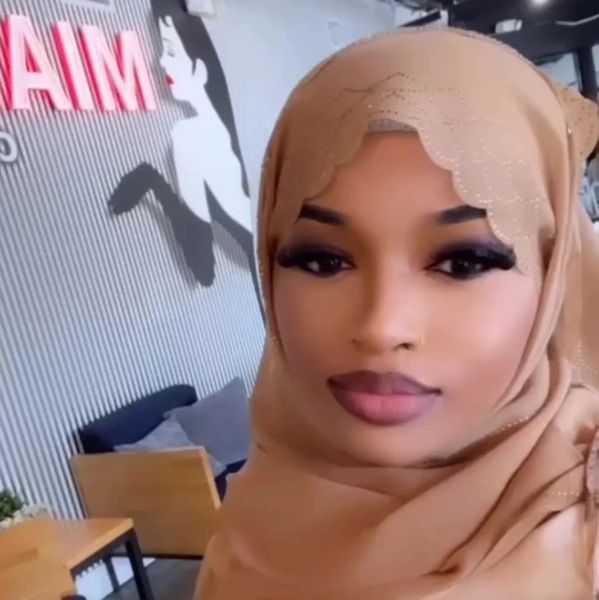 Woman wearing a tan hijab posing indoors near a wall with artwork and neon sign.