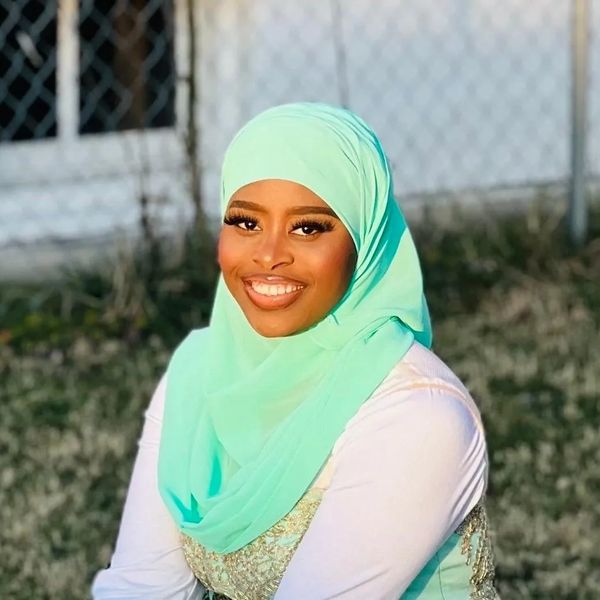 Woman smiling, wearing a light turquoise hijab outdoors.