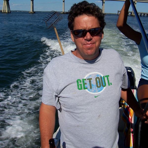 Man wearing sunglasses and gray t-shirt on a boat with a bridge in the background.