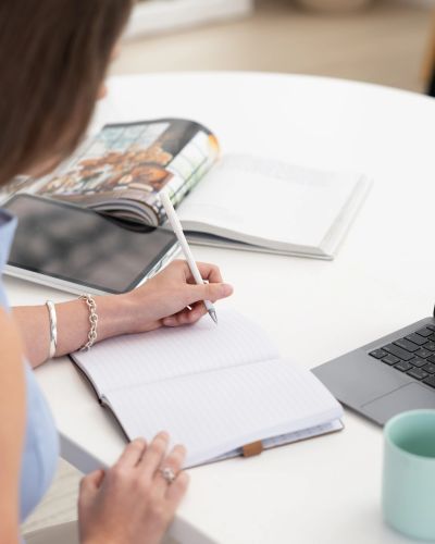 Person writing in a notebook at a desk with a laptop and tablet.