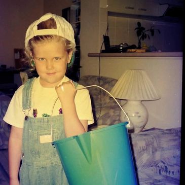 A young child in overalls and a backward cap holding a large green bucket indoors.