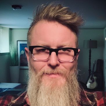 Man with glasses and a long beard indoors, smiling slightly.